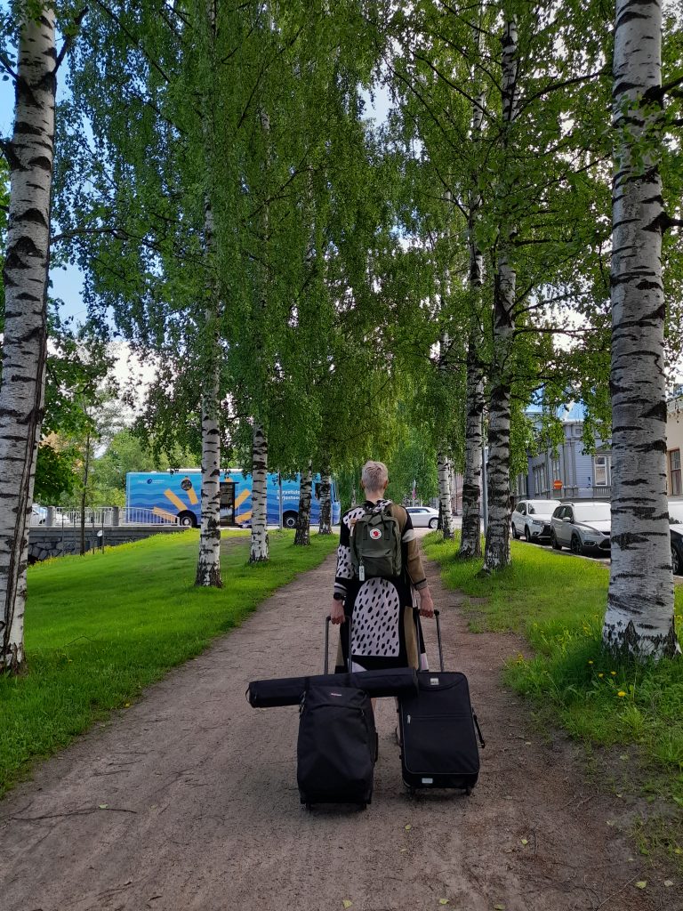 Laura vetää kahta matkalaukkua kohti juna-asemaa. Taustalla näkyy kirjastoauto Kauno.